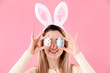 © New Africa - Easter celebration. Smiling woman in headband with bunny ears covering eyes with painted eggs on pink background