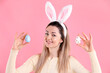 © New Africa - Easter celebration. Portrait of smiling woman in headband with bunny ears holding painted eggs on pink background