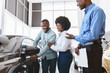 © Prostock-studio - Car sales business. Manager talking to Afro couple, showing them new auto at dealership shop. Young black family selecting vehicle, looking at modern automobile at showroom