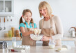 © Prostock-studio - Cooking With Pleasure. Happy little girl in apron mixing dough for cookies or pancakes in glass bowl, cheerful senior grandma helping her grandchild at kitchen. Smiling granny sharing family recipe