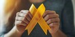© Luisa - Person with dark hair wearing a gray sweater holds yellow support ribbons in both hands against light background.