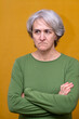 © Manuel - Mature woman with gray hair wearing a green shirt, looking annoyed while standing with crossed arms against a muted orange background