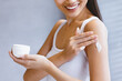 © Prostock-studio - A woman is smiling while she applies cream to her arm with one hand. She holds a small container of cream in her other hand. The setting is bright and indoor, suggesting a skincare routine.