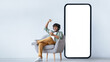 © Prostock-studio - A person sits in a gray chair with a phone in hand, smiling and celebrating. A large blank screen is positioned next to him, highlighting his excitement.