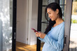 © Wavebreak Media - Mid-adult Asian woman leaning on sliding-door at home in blue top, holding maroon phone, copy space