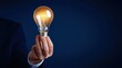 © oksa_studio - Hand of a businessman holding a bright light bulb against a dark blue background, symbolizing ideas, innovation, and success