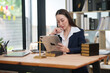 © Thitisak - Asian businesswoman or lawyer in a formal blue suit sitting at a wooden desk, using a digital tablet. The desk is decorated with legal symbols including a gold scale of justice and law books.