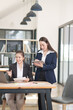 © Thitisak - Two businesswomen in professional suits working together, one holding a laptop and the other taking notes on a tablet, showcasing teamwork and technology in a modern corporate environment.