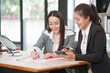 © Thitisak - Two businesswomen in professional suits working together, one holding a laptop and the other taking notes on a tablet, showcasing teamwork and technology in a modern corporate environment.