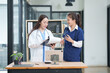 © Thitisak - Two female healthcare workers, one in blue scrubs and another in a white lab coat, are collaborating and reviewing patient charts on a clipboard and tablet. They are engaged in a professional discussi