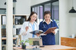 © Thitisak - Two female healthcare workers, one in blue scrubs and another in a white lab coat, are collaborating and reviewing patient charts on a clipboard and tablet. They are engaged in a professional discussi