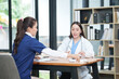 © Thitisak - female doctor in a white lab coat and a medical assistant in blue scrubs are sitting at a wooden desk, collaborating and reviewing medical documents or X-rays. Both are wearing stethoscopes, reflectin