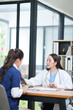 © Thitisak - female doctor in a white lab coat and a medical assistant in blue scrubs are sitting at a wooden desk, collaborating and reviewing medical documents or X-rays. Both are wearing stethoscopes, reflectin