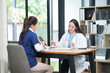 © Thitisak - female doctor in a white lab coat and a medical assistant in blue scrubs are sitting at a wooden desk, collaborating and reviewing medical documents or X-rays. Both are wearing stethoscopes, reflectin