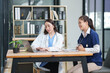 © Thitisak - female doctor in a white lab coat and a medical assistant in blue scrubs are sitting at a wooden desk, collaborating and reviewing medical documents or X-rays. Both are wearing stethoscopes, reflectin