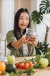 © sofiko14 - A young Asian woman smiles while holding a glass jar filled with nuts, surrounded by fresh fruits and vegetables on a wooden table, suggesting healthy eating and preparation