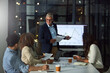 © BuyoutReese06/peopleimages.com - Man, presentation and team in office at night with finance report, data analysis and profit. Mature speaker, staff and document for sales growth, financial graphs and overtime for performance metrics