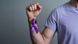 © Rashid - Strong persons arm with a powerful clenched fist displaying a vibrant purple awareness ribbon on their wrist against a simple gray background
