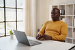 © Tj - Senior Black man using a modern laptop for a video call or online meeting, sitting at a desk in a bright home office, engaging in remote communication