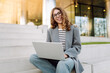 © maxbelchenko - Stylish woman works on laptop while sitting on steps with modern building in background. Beautiful female freelancer in glasses is using a laptop outdoors. Concept: remote work, style, and fashion.