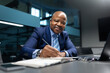 © Anastasiya - Black mature businessman smiling while writing notes at office desk, professional executive lifestyle with focus on leadership, experience, and corporate success.