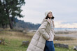 © SHOTPRIME STUDIO - Woman outdoor coat winter fashion in countryside landscape, walking by the coast with beige puffer, jeans and beanie, calm scene with open field and soft light