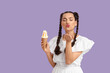 © Pixel-Shot - Beautiful young woman with braids holding tasty ice cream blowing kiss on purple background