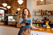 © maxbelchenko - A female barista stands behind the counter, serving freshly brewed coffee. A happy woman in an apron prepares aromatic coffee in a cozy coffee shop. Small business concept, drinks.