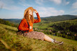© maxbelchenko - Young woman wearing hat and carrying backpack enjoys sunny day on green hill. Traveler feels free as she walks along hiking trail and looks out at mountain landscape. Concept: adventure, relaxation.