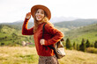 © maxbelchenko - Young woman wearing hat and carrying backpack enjoys sunny day on green hill. Traveler feels free as she walks along hiking trail and looks out at mountain landscape. Concept: adventure, relaxation.