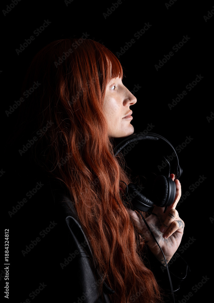 A DJ stands in a dark space, holding headphones while looking to the side. The long red hair contrasts with the background. The person appears to be focused on their music.