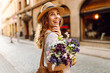 © maxbelchenko - Cute woman strolls along a sunny city street with bouquet of wildflowers. Beautiful tourist in hat enjoys architecture, atmosphere of city with flowers in hands. Concepts of celebration, relaxation