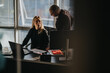 © qunica.com - A professional woman sits at a desk with a laptop, while a man leans over and discusses documents. The modern office setting features blinds, natural light, and work materials.