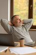 © Daria - Young man with light brown hair is seated at a desk in a modern office, relaxing with a laptop and coffee cup, large windows in the background