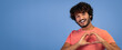 © Prostock-studio - A man stands against a blue background, smiling and forming a heart shape with his hands. He wears a short-sleeved shirt. The scene shows joy and playfulness in his expression.
