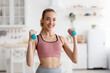 © Prostock-studio - Happy athlete works out at home, exercises for hands and keeps fit during COVID-19 lockdown. Smiling young attractive pretty blonde woman in sportswear lifts dumbbells in minimalist kitchen interior