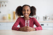 © Prostock-studio - Cheerful african american pretty girl teenager drinking water while sitting at table, smiling at camera, kitchen interior, copy space. Hydration, fresh water drinking for kids, teenagers concept