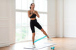 © Prostock-studio - Full And Lower Body Workout. Focused Sporty Woman Exercising With Elastic Rubber Band Lifting And Kicking Leg Aside Standing On Yoga Mat, Listening To Music In Wireless Earpods. Bodyweight Training