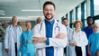 © VAKSMANV - Confident Caucasian male doctor standing in bright corridor crossing arms. Smiling medical leader inspiring diverse team behind him. Showing assurance during busy hospital day. People feeling proud.