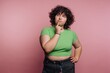 © bodnarphoto - A young person with curly hair wearing a green crop top and black pants, posing thoughtfully against a pink background
