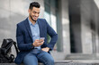© Prostock-studio - Wealthy middle-eastern entrepreneur using cellphone, sitting on the street by office building with backpack and laptop, resting after business event, checking emails, using business app, copy space