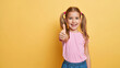 © SerPak - A cheerful girl with pigtails shows a thumbs up gesture against a bright yellow background. Positive attitude and happiness.