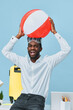 © SHOTPRIME STUDIO - Joyful young man with dark skin and a wide smile holds a colorful beach ball above his head, wearing a white shirt and sunglasses, standing against a light blue background. People lifestyle concept