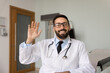 © fizkes - Modern healthcare communication. Screen view portrait smiling young male doctor general practitioner in uniform wave hand hello greet patient on videocall start online telehealth consultation session