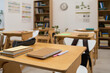 © pressmaster - Empty classroom showing wooden desks with notebooks, pens, and pencil case arranged neatly, bookshelves filled with books in background, educational posters and clock on wall