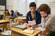 © pressmaster - Two boys sitting at desk reading handmade card with heart drawing, two teenage girls talking in background, classroom setting, school supplies visible