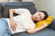 © KMPZZZ - Sick asian young woman in pain, hand holding belly, lying on the sofa with a stomachache, hurt abdomen ache and digestive problem, gastritis or diarrhea. Health care inflammation, abdominal problem