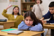 © pressmaster - Caucasian preteen girl sitting at desk with head resting on arms appearing sad, while classmates in background interacting and one boy reaching toward her in classroom setting