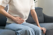 © KMPZZZ - Sick asian young woman in pain, hand holding belly, sitting on sofa with a stomachache, hurt abdomen ache and digestive problem, gastritis or diarrhea. Health care inflammation, abdominal problem.