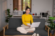© Wasana - Young asian woman working remotely from home office using laptop on sofa